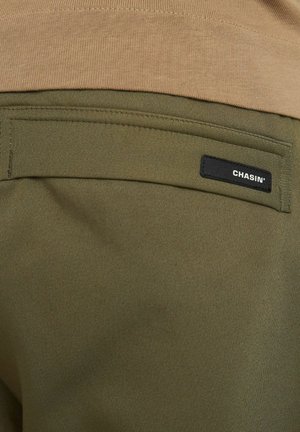 Olijfgroene broek met een gladde textuur, voorzien van een achterzak met een zwart stoffen label waarop "CHASIN'" staat. Het bovenste gedeelte is tan.