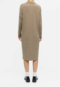 Personne portant une robe longue beige côtelée en tricot à manches longues, des chaussettes blanches et des chaussures noires, debout de dos sur un fond blanc.