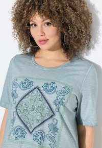 Camiseta de manga corta en azul claro con un intrincado estampado de paisley en verde y azul marino, detalles en forma de diamante en el centro y una tela suave al tacto.
