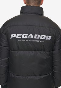 Giacca a vento nera con colletto alto e polsini elasticizzati. Logo "PEGADOR" in bianco sul retro, insieme a "CERTIFIED AUTHENTIC STREETWEAR."