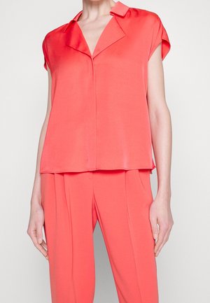 Blusa de seda coral con mangas cortas y escote en V. Presenta un corte relajado y líneas limpias, complementando los pantalones coral a juego que lleva debajo.