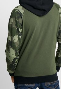 Bluza z kapturem w czarnym kolorze kaptura, oliwkowo-zielonym korpusie i rękawach w kamuflażowy wzór. Miękka tkanina z ściągaczami na mankietach i dole.