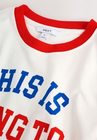Maglietta bianca a maniche corte con colletto a costine rosso, con testo audace in blu e rosso che recita "QUESTO STA PER". Materiale in cotone.