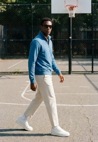 Mann mit Sonnenbrille, blauem Pullover und weißen Hosen geht auf einem Freiluft-Basketballplatz nahe dem Korb und einem Maschendrahtzaun.