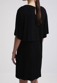 Černé šaty s volnými křídly, které mají otvor na zadní straně. Hladká tkanina, délka po kolena a uvolněná silueta.