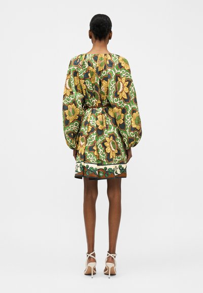 Γυναίκα με κοντό λουλουδάτο φόρεμα με πράσινα, κίτρινα και καφέ σχέδια, μακριά φουσκωτά μανίκια και λευκά σανδάλια με ψηλά τακούνια, γυρισμένη προς τα πίσω.