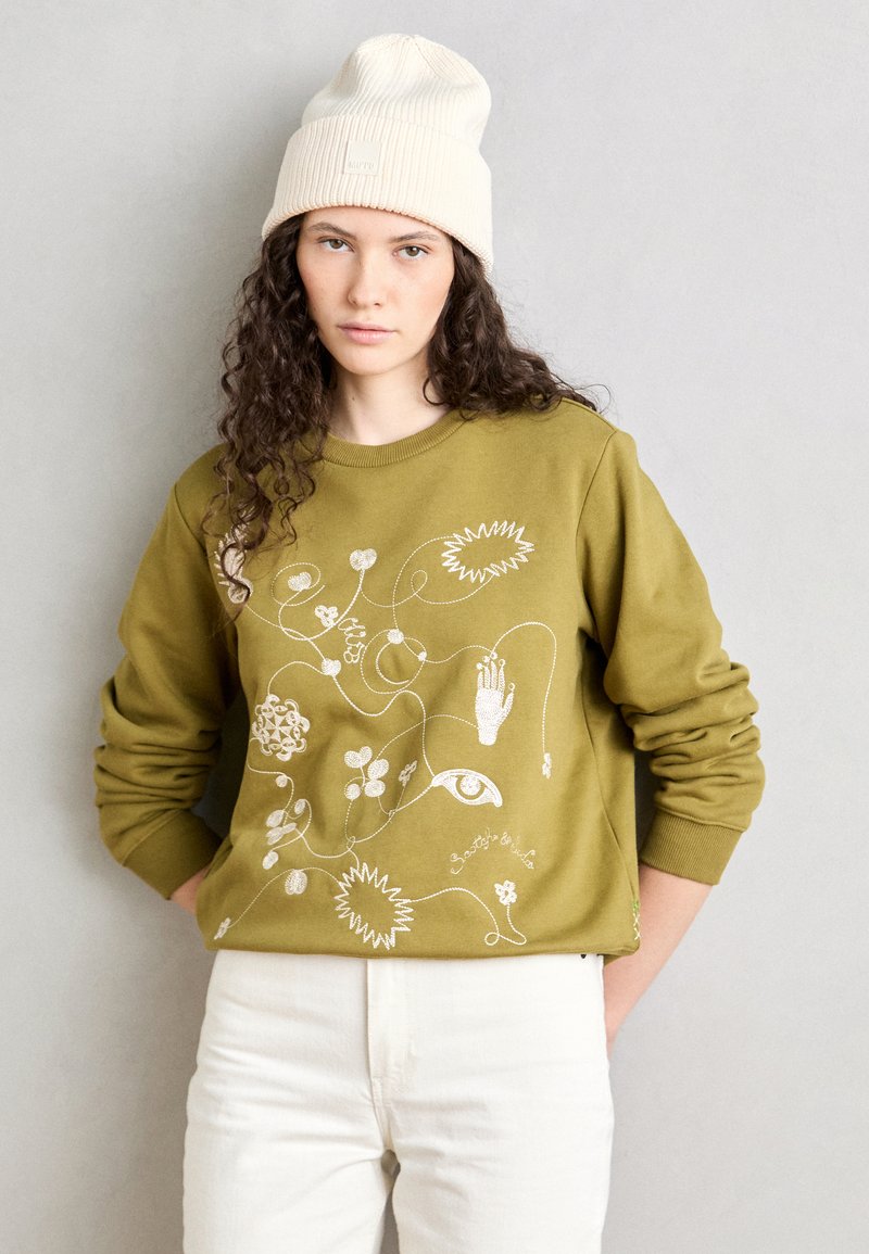 Sweatshirt vert olive avec des motifs floraux et abstraits brodés en blanc. Associé à un pantalon crème et un bonnet côtelé blanc cassé.