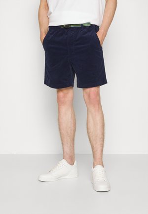 Granatowe sztruksowe szorty z elastycznym paskiem w talii i bocznymi kieszeniami, zestawione z zielono-paskowanym paskiem i białymi trampkami.