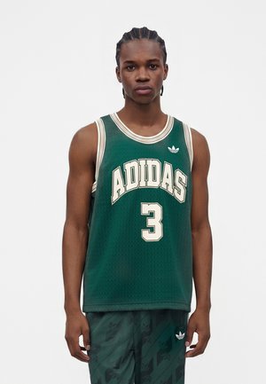 Mladý muž s vrkočmi, ktorý nosí tmavozelený basketbalový dres Adidas s číslom tri a zladené šortky na jednofarebnom pozadí.