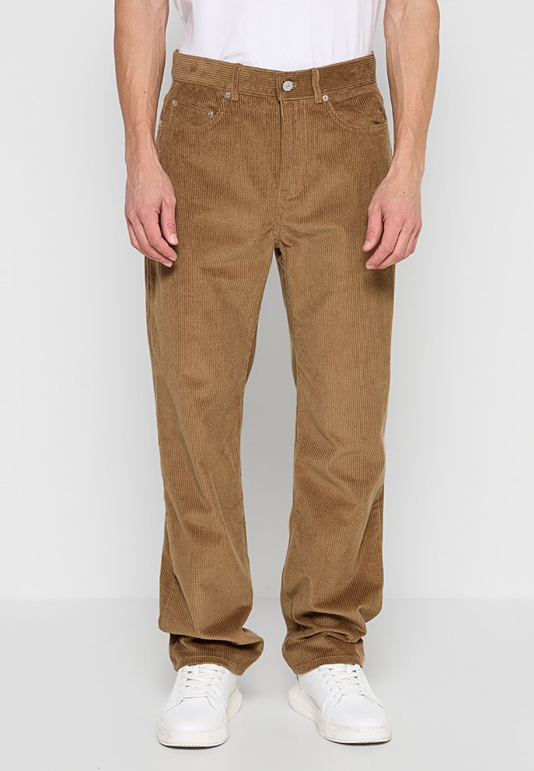Pantalon en velours côtelé marron avec une coupe droite, doté d'une fermeture à bouton et de poches latérales, porté avec des baskets blanches.