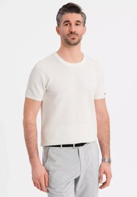 Suéter de punto blanco de manga corta con cuello redondo, con un patrón texturizado y un sutil acento del logotipo en la manga, combinado con pantalones claros.