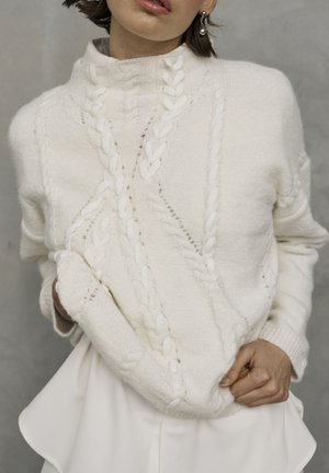 White knitted sweater with a high collar and textured cable patterns. Fabric appears soft, with ribbed cuffs and a relaxed fit.