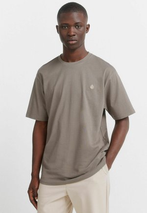 Młody mężczyzna o krótkich włosach, ubrany w prostą taupe koszulkę i jasnobeżowe spodnie, stojący z jedną ręką w kieszeni na białym tle.