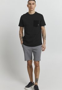 Camiseta de algodón negra con un bolsillo en el pecho izquierdo, combinada con pantalones cortos grises. El modelo lleva zapatillas negras, que presentan un diseño simple y casual.