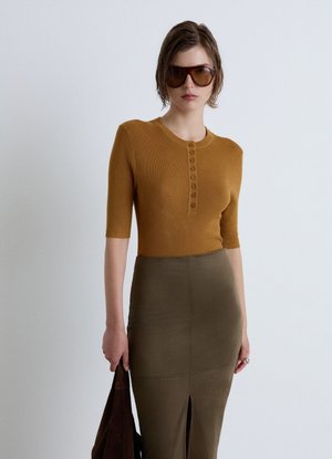 Woman wearing amber sunglasses, a mustard ribbed buttoned short-sleeve top, and an olive high-waisted skirt holding a dark brown bag.