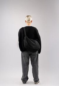 Borsa a tracolla nera realizzata in tessuto morbido, con una forma curva e tracolla regolabile, indossata in modo informale con pantaloni grigi morbidi e un maglione.