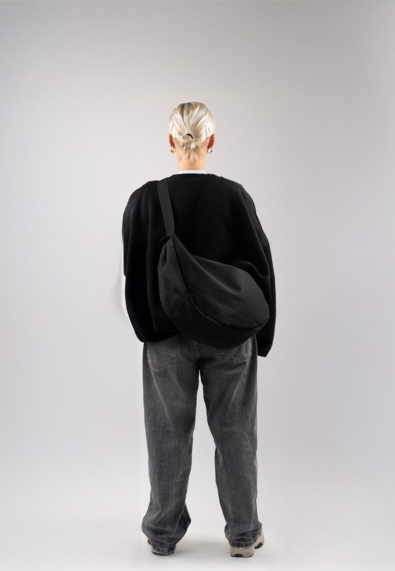 Borsa a tracolla nera realizzata in tessuto morbido, con una forma curva e tracolla regolabile, indossata in modo informale con pantaloni grigi morbidi e un maglione.