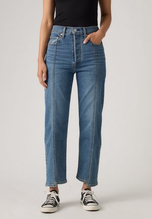Person trägt hochtaillierte blaue Jeans mit zentralen vorderen Nähten und schwarz-weißen Sneakern, eine Hand in der Tasche, steht vor neutralem Hintergrund.