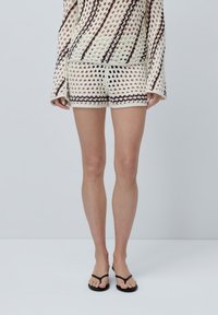 Maglione a maniche lunghe con scollatura ampia, di colore crema con strisce marroni. Abbinato a pantaloni corti crema coordinati. Infradito nere.