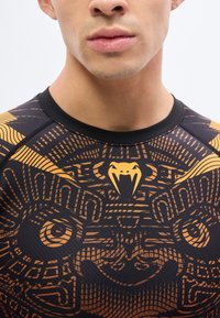 Νέος άνδρας που φοράει αθλητική μπλούζα σε μαύρο και πορτοκαλί χρώμα με μια λεπτομερή αφηρημένη εικόνα προσώπου λέοντα και ένα κίτρινο λογότυπο κεφαλής φιδιού στο στήθος.