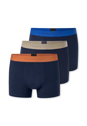 Hemdloser Mann mit kurzen braunen Haaren, der marineblaue Boxershorts mit blauem Bund trägt und nach unten schaut.