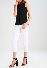 Zwarte mouwloze top met een V-hals, gecombineerd met witte cropped pants en zwarte hakken met enkelbandjes, tegen een neutrale achtergrond.