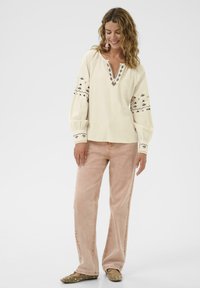 Femme aux cheveux bouclés portant une blouse longue à manches brodées crème, un jean rose clair et des ballerines marron à clous, souriant les yeux fermés.