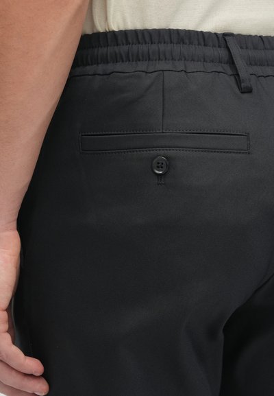 Calças pretas com um bolso traseiro plano com um único botão, faixa elástica na cintura e textura de tecido suave.