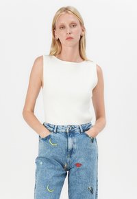 Witte ribgebreide mouwloze top met ronde halslijn, gecombineerd met lichtblauwe high-waisted denim jeans met kleurrijke geborduurde ontwerpen.