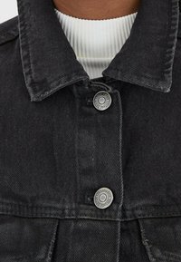 Černá džínová bunda s špičatým límcem, stříbrným uzávěrem na knoflíky a na krku jsou viditelné bílé ribbed vrstvy.