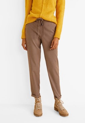 Vrouw draagt bruine broek met trekkoord, opgerolde zomen, lichtbruine veterlaarzen en een geel gebreide trui, staand tegen een witte achtergrond.