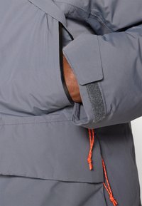 Pelēks izolēts parka, kas aprīkots ar regulējamu auklu, ar rāvējslēdzēju aizveramu kabatu un ar āķi- un cilpas tipa aproču aizdari. Audums ar faktūru un oranžiem akcentiem.