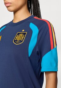 Κοντινό πλάνο ενός ατόμου που φοράει σπορ μπλούζα ναυτικού μπλε χρώματος με το σήμα της ποδοσφαιρικής ομάδας της Ισπανίας, και λεπτομέρειες στους ώμους σε ανοιχτό μπλε και κόκκινο-κίτρινο ριγέ σχέδιο.