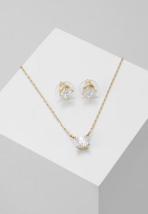 Gold chain necklace and matching stud earrings with square clear gemstones on a white cube against a gray background.