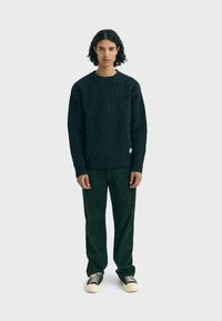 Dark ribbed sweater with crew neckline paired with green corduroy pants and black sneakers. Model stands against a light background.