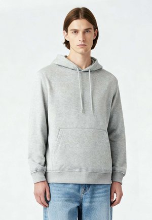 Giovane uomo con capelli castani lisci che indossa una felpa con cappuccio grigio chiaro e jeans blu, in piedi davanti a uno sfondo chiaro e semplice.