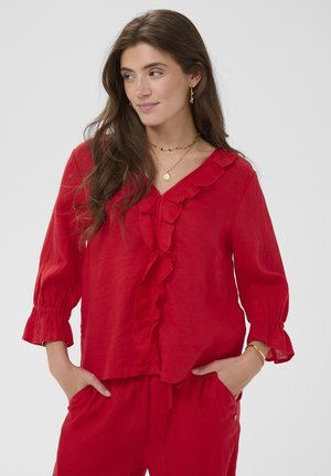 Kobieta o długich brązowych włosach, ubrana w czerwoną bluzkę z falbanami i dopasowane spodnie, ręce w kieszeniach, patrząca na bok z delikatnym uśmiechem.