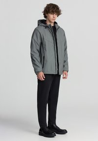 Giacca impermeabile grigia con cappuccio, chiusura con zip e tasche laterali, abbinata a jeans neri e stivali neri con lacci.