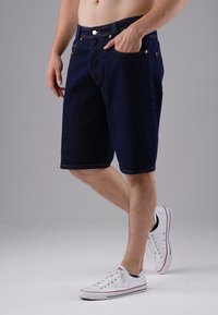 Dark blue denim shorts with a straight cut, five pockets, and contrasting yellow stitching. Paired with white sneakers.
