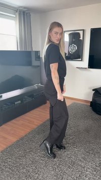 Fiatal nő hosszú szőke hajjal, fekete ruhában és csizmában, szürke szőnyegen áll a nappaliban, ahol TV és keretezett műalkotás látható.