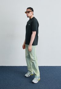 Black cotton t-shirt with logo, light green denim pants, and silver athletic shoes with yellow accents; person wearing brown sunglasses, standing sideways.