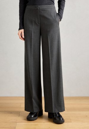 Grijze, wijde broek met een gestructureerd patroon, hoge taille en zijzakken, gecombineerd met zwarte platform schoenen op een houten vloer.