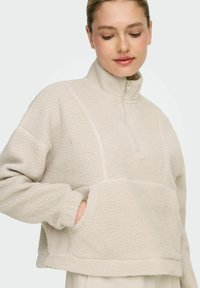 Fleece pullover in light beige with a quarter-zip, textured surface, drop shoulders, and side pockets. Features stitched panel accents.