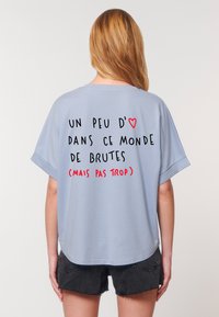 A person faces away, wearing a light blue t-shirt with black and red text saying, "Un peu d'♥ dans ce monde de brutes (mais pas trop)."