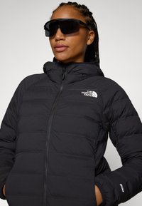 Svart puffjacka med dragkedja fram och huva. Har horisontella sömmar, North Face-logotyp och elastiska ärmslut. Bärs med kraftiga svarta solglasögon.