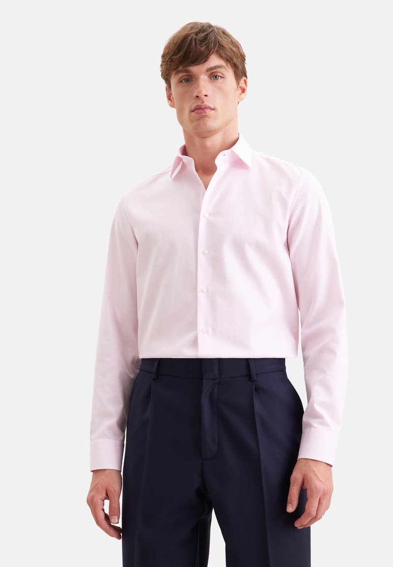 Homme portant une chemise rose clair boutonnée rentrée dans un pantalon tailleur bleu marine foncé sur un fond blanc uni.