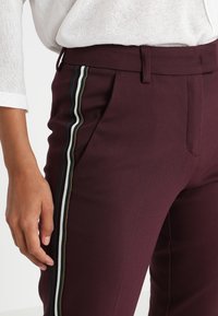Bordowe spodnie klasyczne charakteryzujące się slim fit, bocznymi paskami w kolorach zielonym, białym i czarnym, o gładkiej fakturze i bocznych kieszeniach.