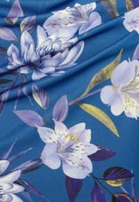 Sininen kangas, jossa on suuria, yksityiskohtaisia violetteja ja valkoisia kukkakuvioita vihreiden lehtien kera, luoden eloisan ja tekstuurisen ilmeen.