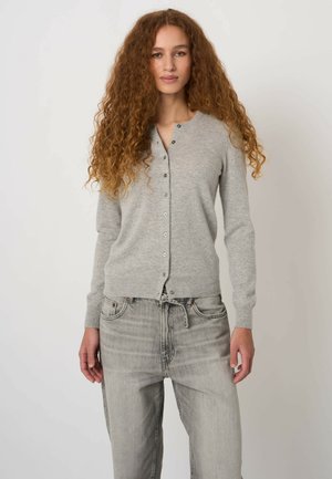 Kobieta o długich kręconych włosach, ubrana w jasno szary, zapinany na guziki kardigan oraz wyblakłe szare jeansy na jednolitym tle.