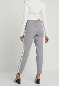 Grijze tapse broek met witte zijsstripes. Voorzien van een elastische tailleband en achterzakken. Draagt in combinatie met zwart-witte gestreepte hakken.